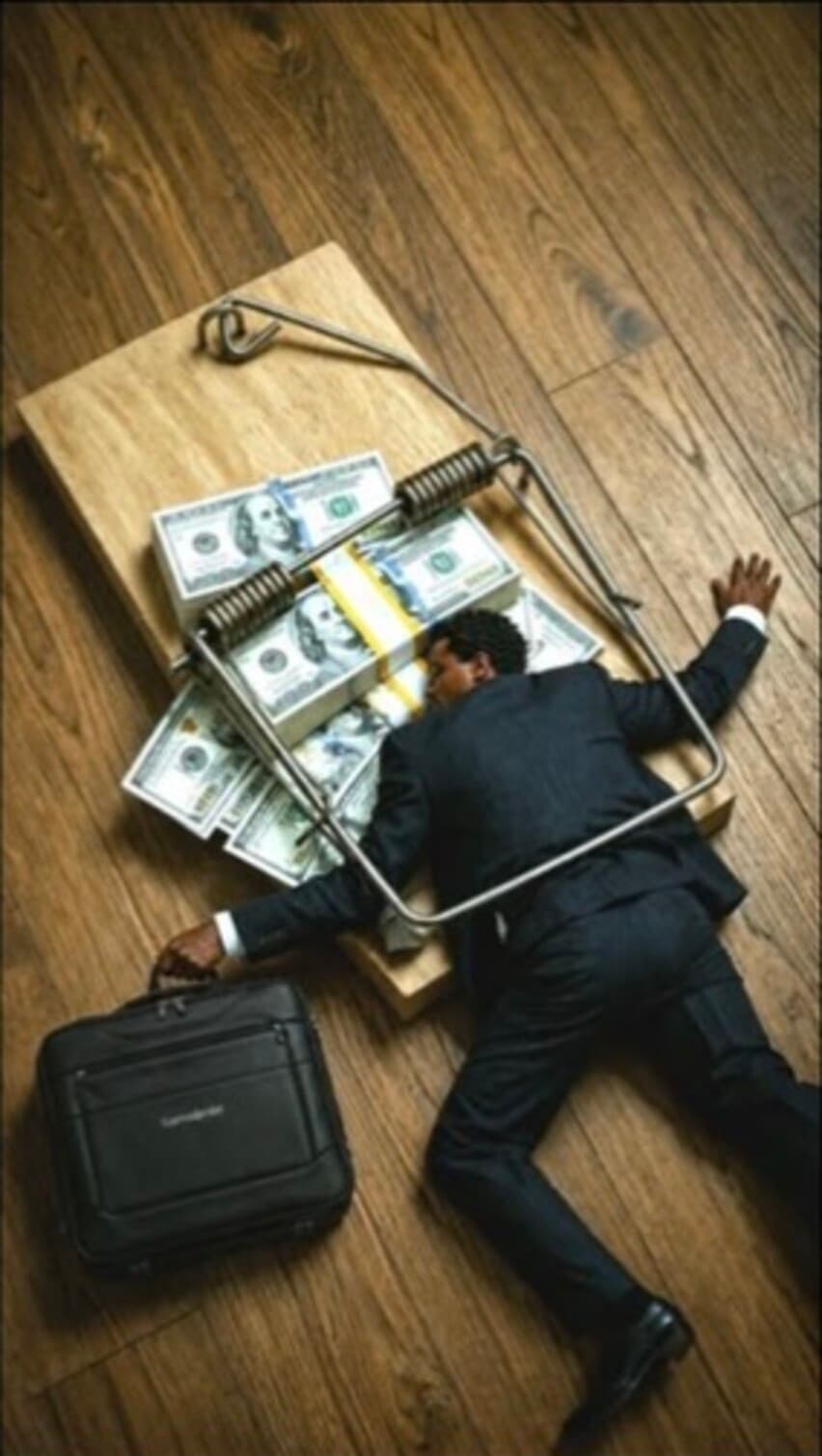 A man in a business suit caught in a mousetrap baited with bundles of money.